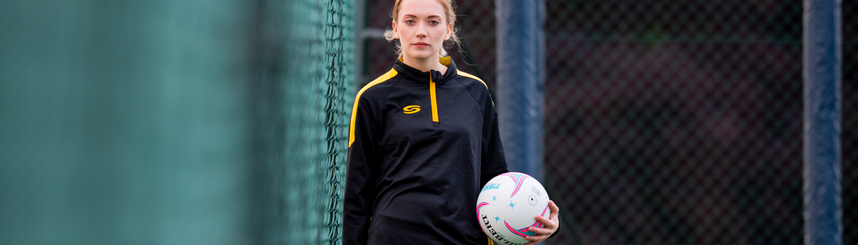 Desktop campaign slider for our netball teamwear solution Netball player holding a ball wearing black/yellow core midlayer